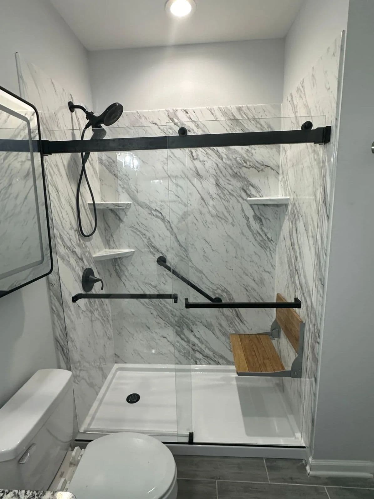 A walk-in shower with white and gray marbled walls, sliding glass doors, black fixtures, and a folding shower bench seat.
